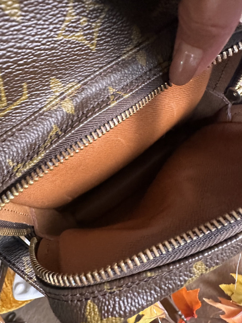 Close-up of a brown monogram handbag interior with a gold zipper and a tan lining, shown with a finger pointing into the compartment.