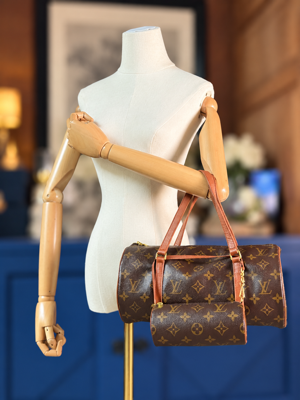 A Louis Vuitton monogram duffle bag in brown and tan is displayed on a white dress form mannequin with wooden arms, set against a softly blurred indoor background with blue paneling.